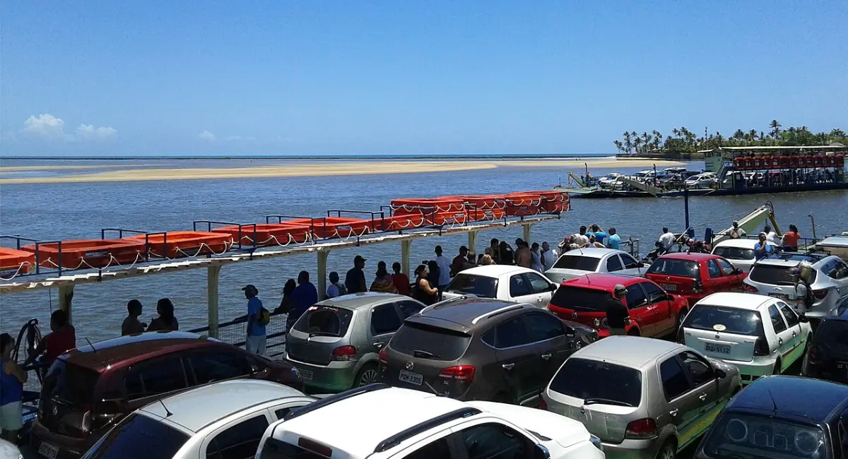 Travessia de balsa entre Porto Seguro e Arraial d'Ajuda, mostrando carros e passageiros a bordo, com outra balsa cruzando o Rio Buranhém ao fundo sob céu limpo.