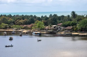 Vista aérea da charmosa Vila de Caraíva na Bahia, mostrando o encontro do rio (Rio Caraíva) em primeiro plano com o mar azul no horizonte. Na margem do rio, é possível ver casas rústicas, muita vegetação tropical, e pequenas canoas e barcos de pesca atracados ou navegando.