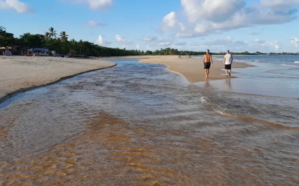 Riacho Barramares encontra o mar na Praia de Taperapuã, Porto Seguro, com dois homens caminhando na areia dourada sob um céu azul.