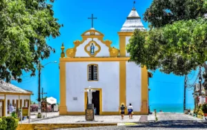 Igreja Nossa Senhora d'Ajuda em Arraial d'Ajuda, Bahia, com vista para o mar e árvores verdes.