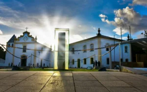 O Marco do Descobrimento em frente à Igreja Nossa Senhora da Pena, na Cidade Histórica de Porto Seguro, com o sol ao fundo.