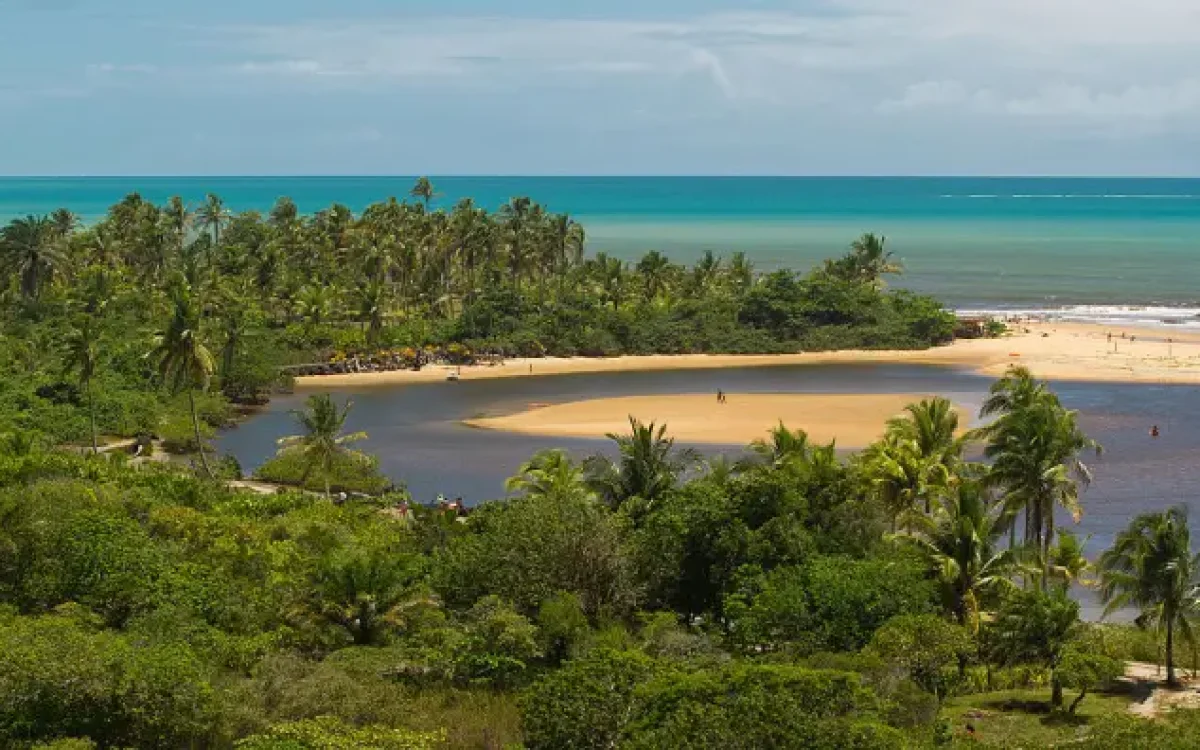 Uma vista aérea que mostra exatamente onde fica Caraíva: no encontro mágico do Rio Caraíva com o oceano, em meio a uma exuberante vegetação tropical e um mar azul-turquesa.