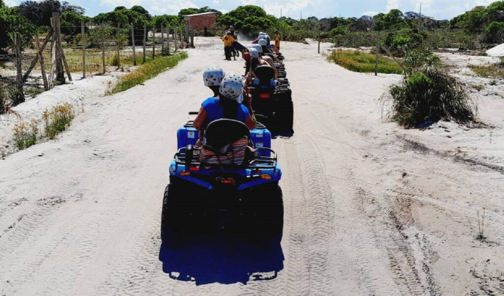 Grupo em passeio de quadriciclo em Porto Seguro, seguindo em fila por trilha de terra na Orla Norte, com vegetação e céu azul ao fundo.