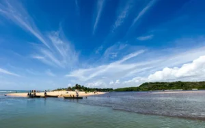 Vista aérea do encontro do Rio Caraíva com o mar, revelando as praias de Caraíva com coqueiros e canoas, sob um céu azul vibrante com nuvens finas.
