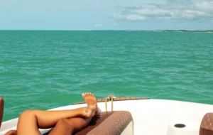 Pés de uma pessoa relaxando e bronzeando em um deck de lancha luxuosa com mar azul-turquesa de fundo, sugerindo um Passeio de Lancha Caraíva Praia do Espelho: Privativo.