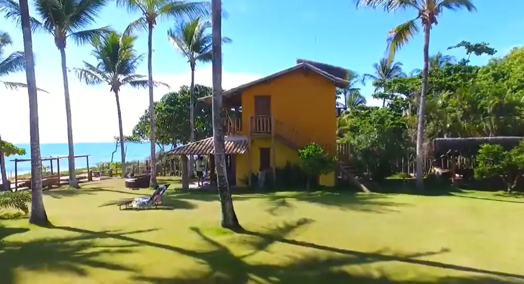 Fachada rústica amarela da Pousada Cares de Mar cercada por coqueiros e gramado verde, com vista para o mar azul ao fundo sob céu limpo.