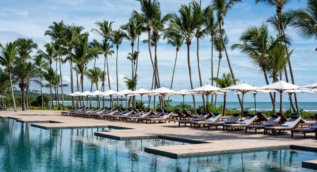 Área da piscina de borda infinita do Hotel Fasano Trancoso, cercada por espreguiçadeiras com ombrelones brancos e coqueiros altos frente ao mar.