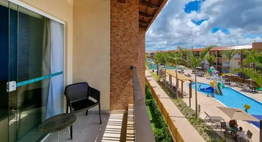 Vista da varanda de um quarto no Ondas Praia Resort em Porto Seguro, mostrando a área de lazer com piscinas infantis, espreguiçadeiras e palmeiras sob céu nublado.
