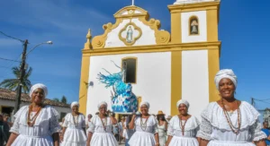 Mulheres vestidas de baianas tradicionais carregando um andor azul com a imagem de Iemanjá em frente à Igreja de Nossa Senhora d'Ajuda, em Arraial d'Ajuda.