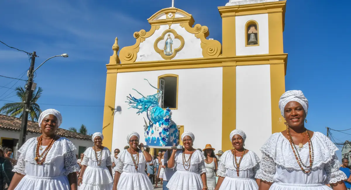 Mulheres vestidas de baianas tradicionais carregando um andor azul com a imagem de Iemanjá em frente à Igreja de Nossa Senhora d'Ajuda, em Arraial d'Ajuda.