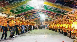 Apresentação de quadrilha junina com dançarinos em trajes coloridos sob teto decorado com bandeirolas na Festa de São João de Porto Seguro.