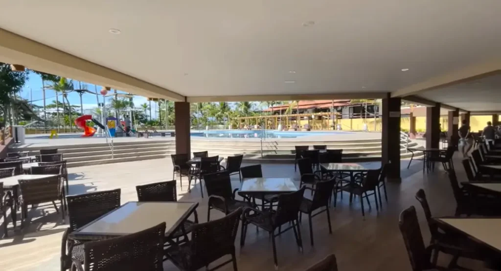 Área de lazer coberta do Porto Seguro Praia Resort com mesas e cadeiras, vista para a piscina e parque aquático infantil ao fundo.