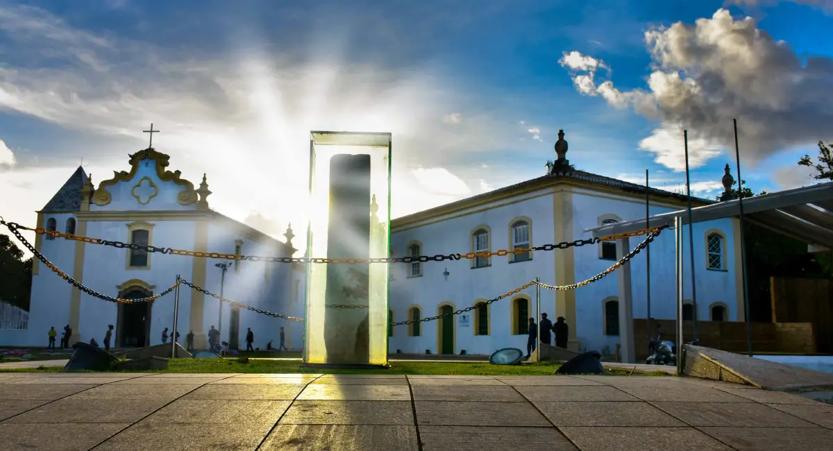 Marco do Descobrimento protegido por redoma de vidro no Centro Histórico de Porto Seguro, Bahia, com a Igreja de Nossa Senhora da Pena ao fundo e raios de sol ao entardecer.