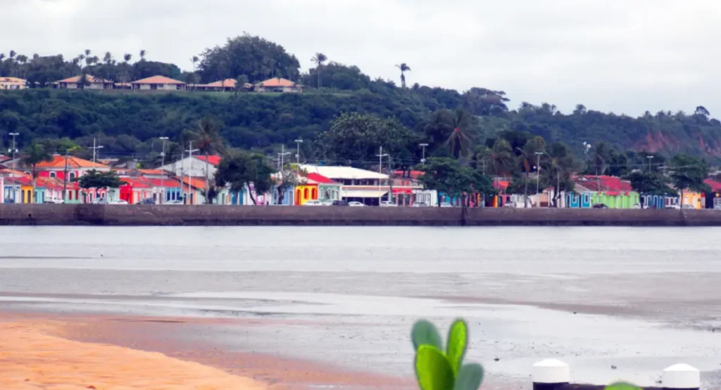Vista panorâmica diurna da Passarela do Alccol em Porto Seguro, com casario colorido histórico à beira do Rio Buranhém e vegetação ao fundo.