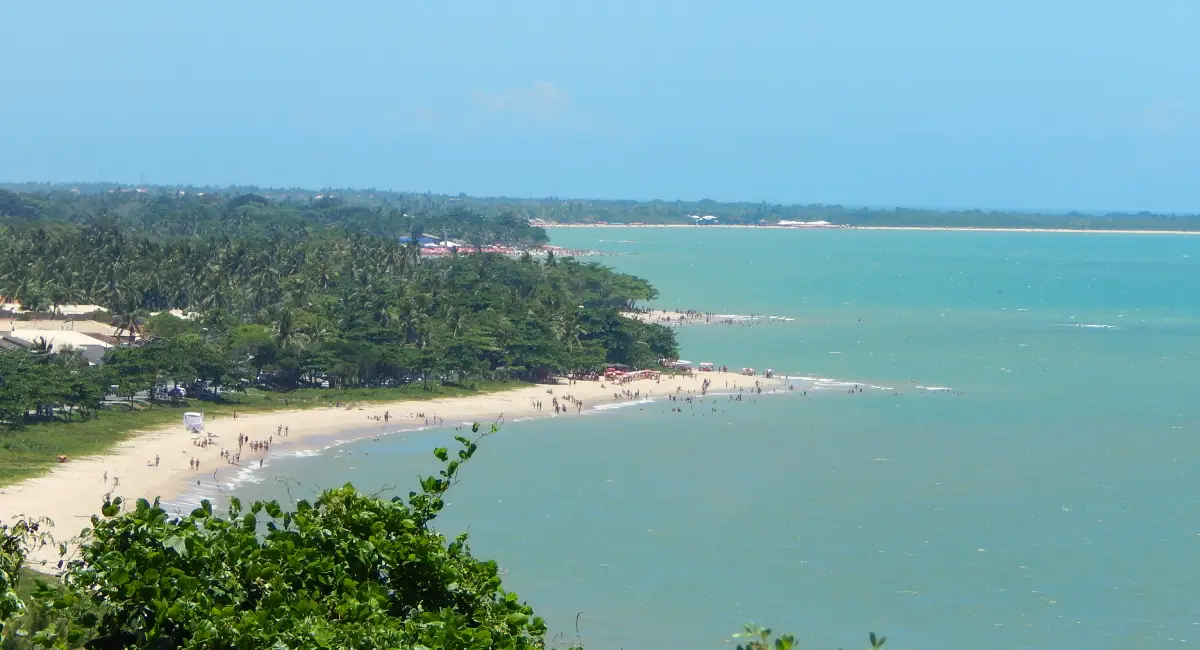 Vista panorâmica a partir do mirante da Cidade Histórica de Porto Seguro, Bahia, exibindo a Orla Norte com praias de águas esverdeadas, vegetação costeira abundante e banhistas na faixa de areia sob um céu azul claro.