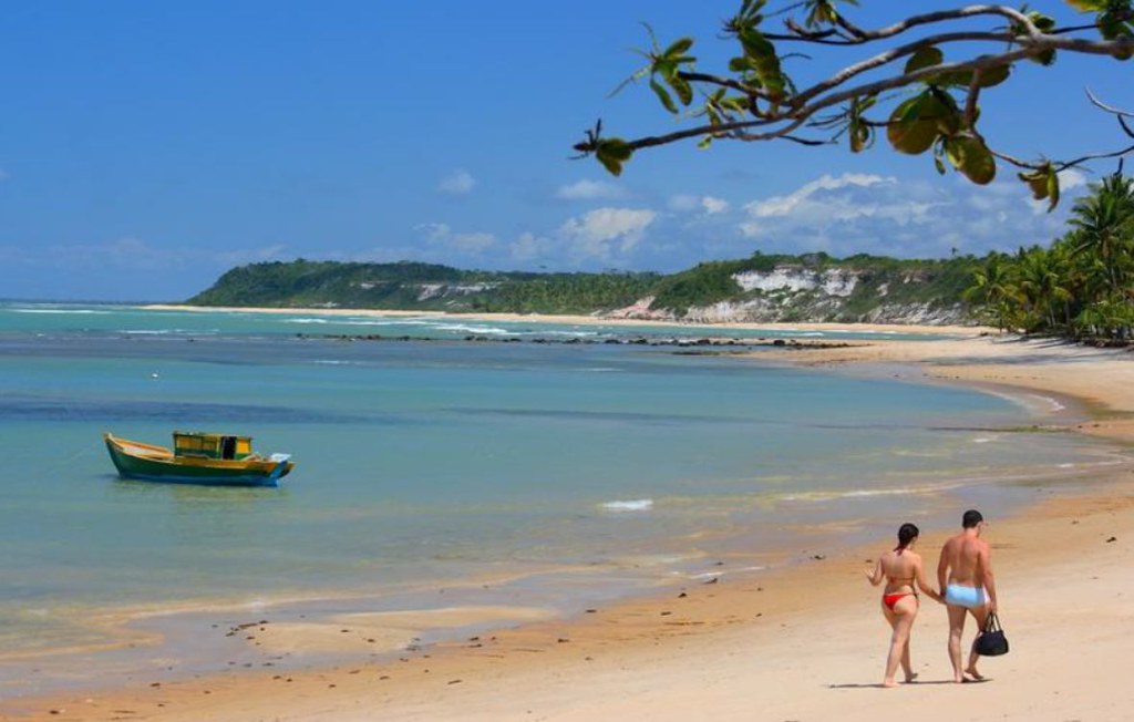 Vista panorâmica da Praia do Espelho na Bahia, mostrando um casal caminhando na areia, um barco de pesca no mar calmo e falésias brancas ao fundo sob céu azul.