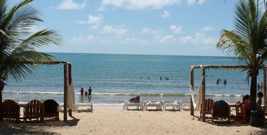 Vista da Praia do Mutá em Porto Seguro com cadeiras de sol na areia, coqueiros e mar calmo com banhistas ao fundo sob céu azul.
