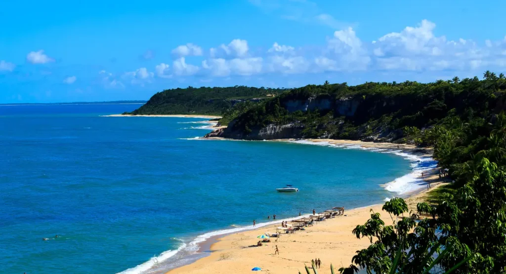 Vista panorâmica da Praia dos Nativos em Trancoso, Bahia, exibindo a longa faixa de areia dourada, mar em tons de azul e verde, falésias cobertas por vegetação nativa ao fundo e banhistas aproveitando o dia sob sol forte.