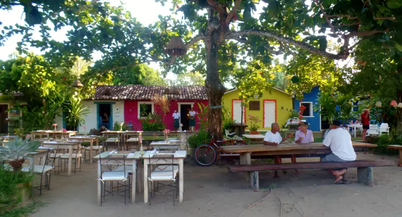 Vista do rústico e charmoso Quadrado de Trancoso, Bahia, sob uma grande árvore. Pessoas estão sentadas em bancos de madeira, e mesas de restaurante estão dispostas na areia. Ao fundo, casinhas históricas e coloridas com portas e janelas simples completam o cenário. O chão é de terra batida e a atmosfera é relaxante.