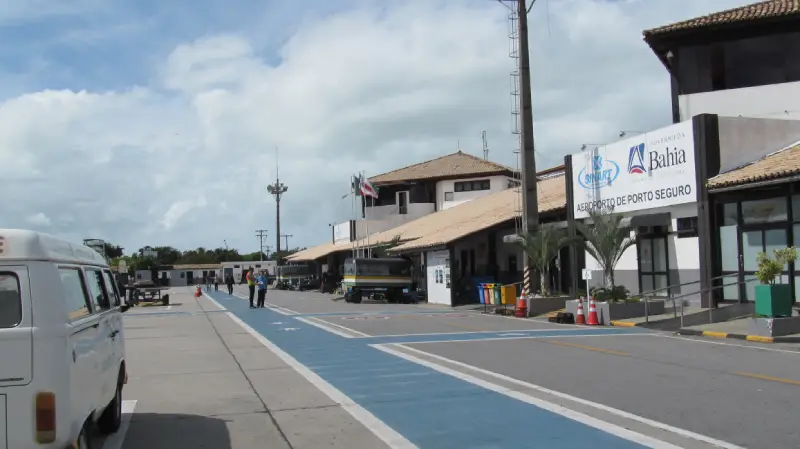 "Fachada externa branca e marrom do Aeroporto de Porto Seguro em um dia nublado. Uma van branca está estacionada à esquerda e um caminho azul claro leva à entrada."