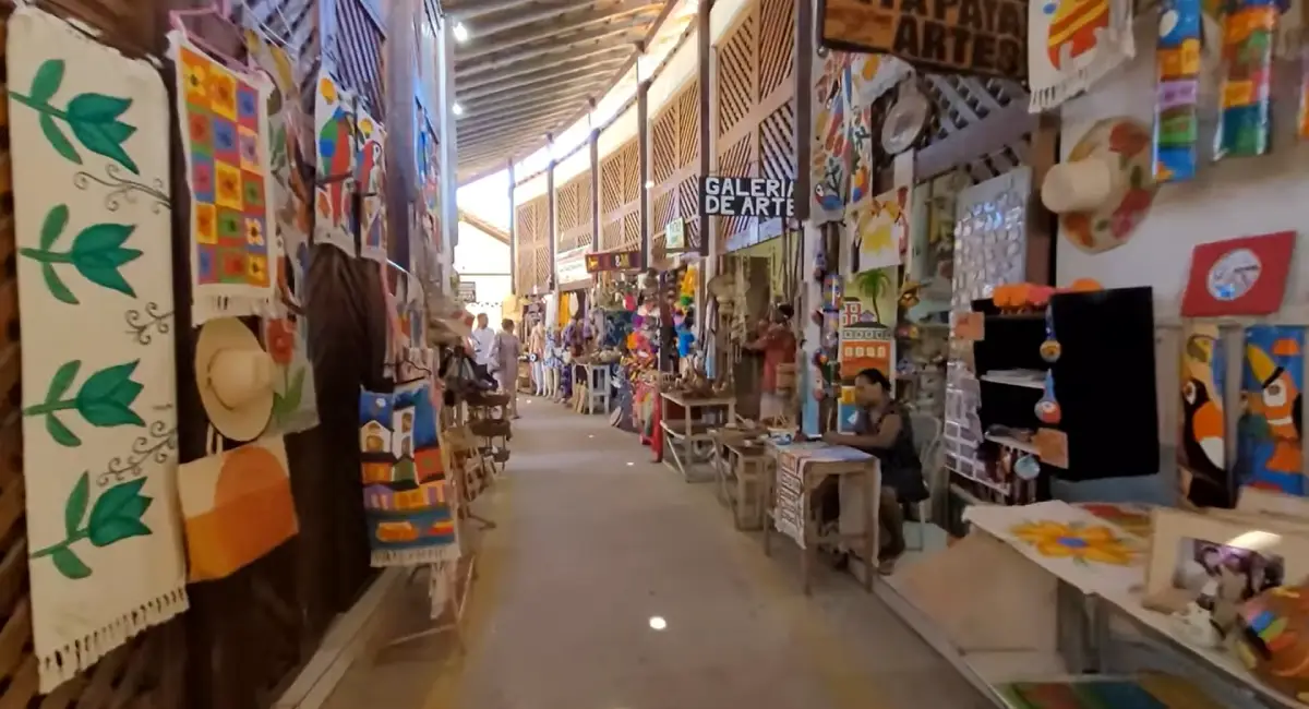 Corredor interno de um mercado de artesanato na Costa do Descobrimento, com diversas lojas exibindo quadros coloridos, chapéus, bolsas e arte indígena local.