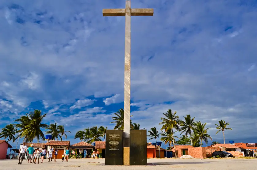 Monumento do Cruzeiro em Coroa Vermelha, símbolo da primeira missa celebrada no Brasil, com turistas caminhando ao redor sob céu com nuvens e coqueiros ao fundo.