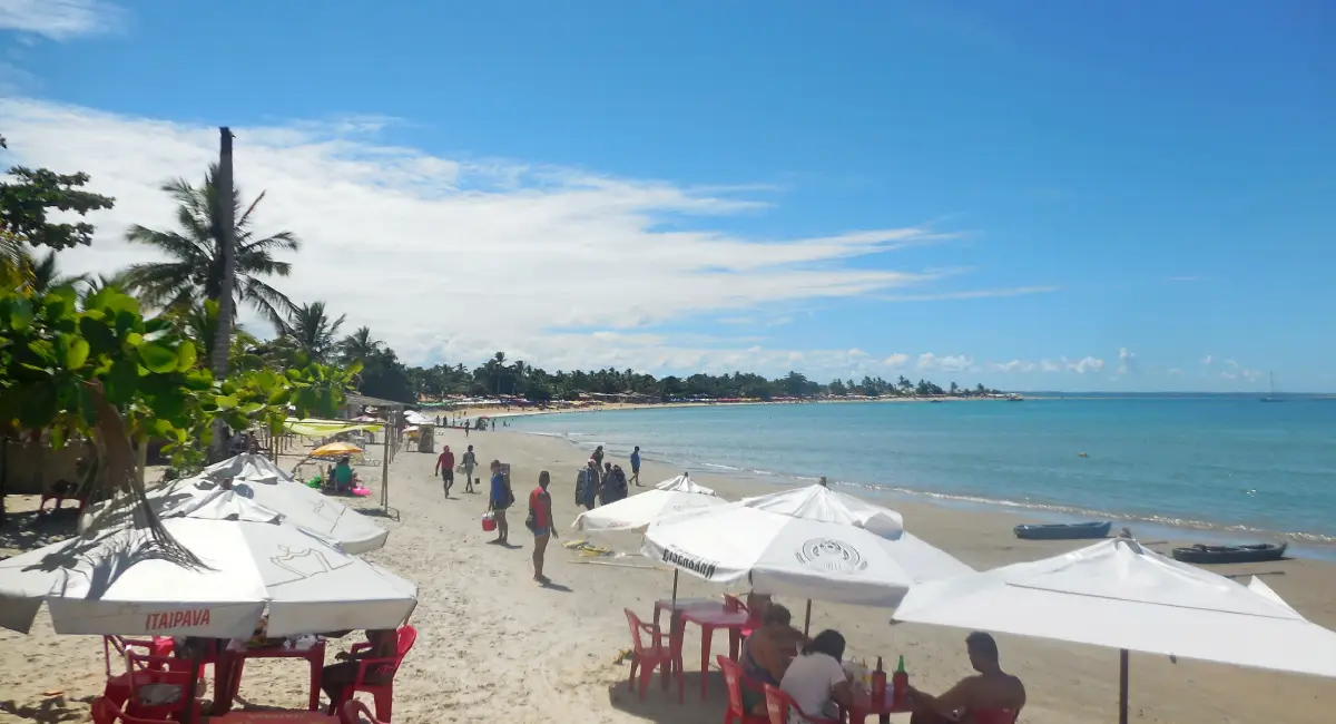 Vista da Praia de Coroa Vermelha em Santa Cruz Cabrália, com mesas e guarda-sóis brancos na areia, banhistas caminhando e o mar calmo onde se forma o Caminho de Moisés.