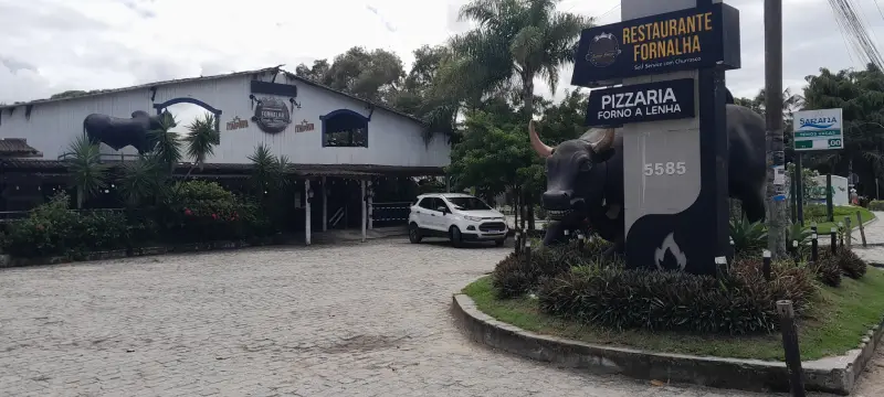 Fachada do Restaurante Fornalha na Orla Norte de Porto Seguro, destacando o totem de entrada com indicações de pizzaria forno a lenha e self-service com churrasco.