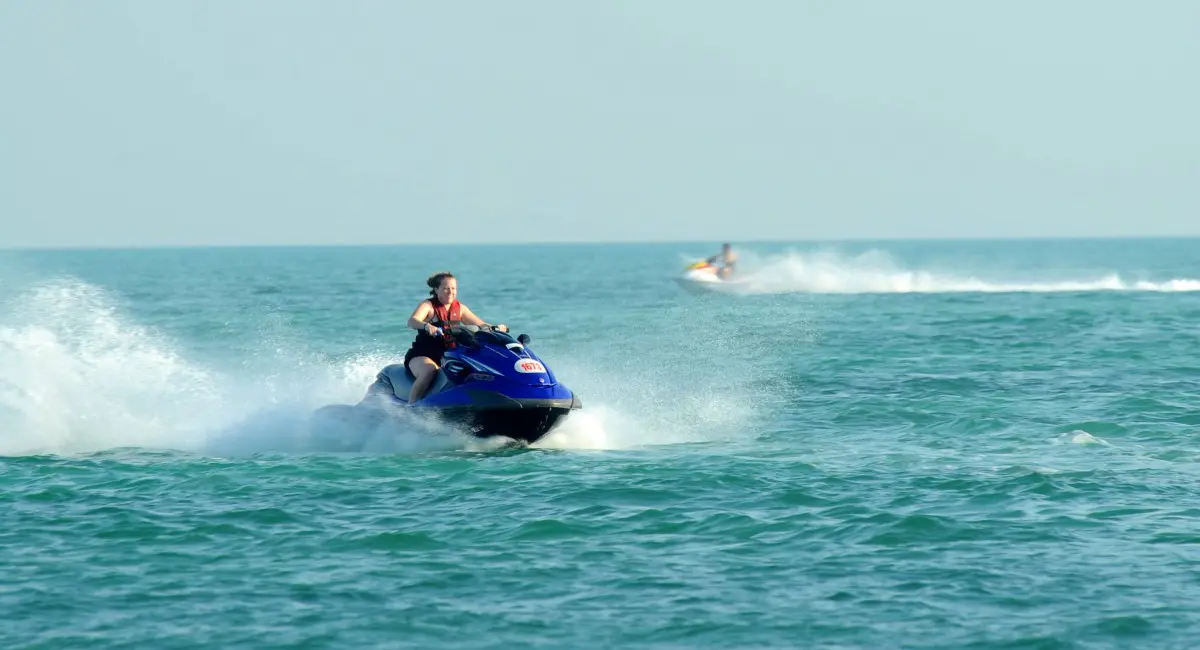 Mulher pilotando um jet ski azul em alta velocidade nas águas calmas e esverdeadas da Praia de Taperapuã, em Porto Seguro, Bahia.