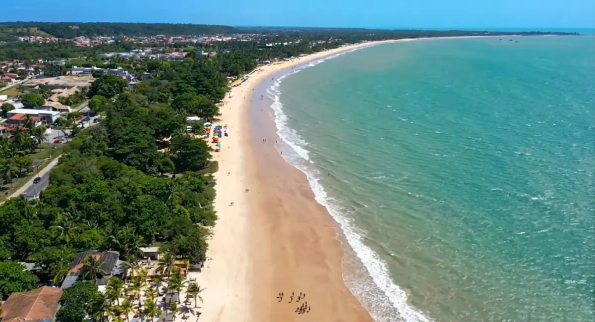 Vista aérea panorâmica da Praia de Taperapuã em Porto Seguro, Bahia. À esquerda, a orla urbanizada com hotéis, pousadas, casas de veraneio e barracas de praia famosas integradas à vegetação nativa densa. Ao centro, a longa faixa de areia dourada com turistas caminhando e jogando futebol. À direita, o mar de cor azul-turquesa cristalino com pequenas ondas brancas quebradas na areia. Ao fundo, no horizonte, o centro urbano de Porto Seguro. Imagem focada em mostrar a infraestrutura hoteleira e a beleza natural da orla de Taperapuã para opções de hospedagem.