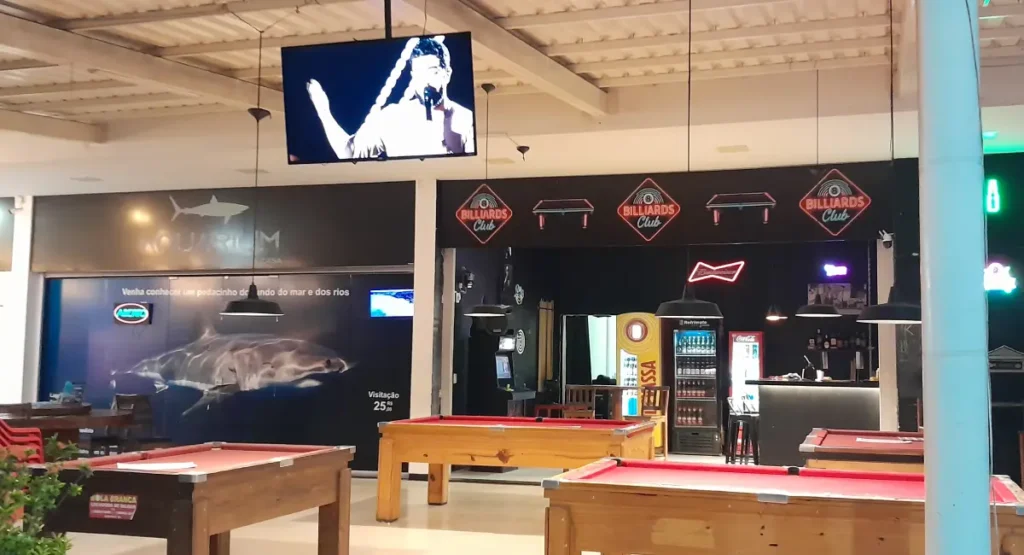 Interior de um complexo de lazer na Praia de Taperapuã com mesas de sinuca vermelhas, bar ao fundo e decoração temática com um grande painel de tubarão e telões.