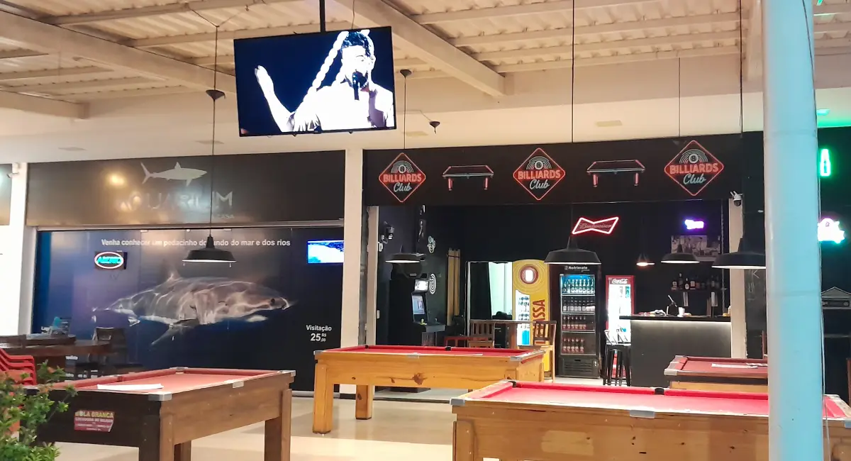 Interior de um complexo de lazer na Praia de Taperapuã com mesas de sinuca vermelhas, bar ao fundo e decoração temática com um grande painel de tubarão e telões.