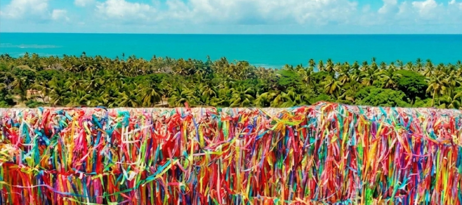 Milhares de fitinhas coloridas de "Lembrança de Arraial d'Ajuda" amarradas no mirante atrás da Igreja Matriz de Nossa Senhora d'Ajuda. Ao fundo, vista panorâmica do mar azul turquesa e coqueirais sob um céu com nuvens brancas na Costa do Descobrimento, Bahia. Fitinhas que Conectam