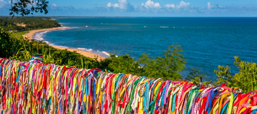 Mirante do Quadrado de Trancoso com centenas de fitinhas coloridas de "Lembrança do Senhor do Bonfim" amarradas na mureta. Ao fundo, vista panorâmica da Praia dos Coqueiros e do mar da Bahia sob um céu com nuvens, destacando a natureza exuberante da Costa do Descobrimento. Fitinhas que Conectam