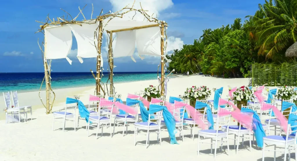 Decoração de festa à beira-mar com altar rústico de madeira e tecidos brancos, cadeiras decoradas com laços azuis e rosa em praia de areia branca, ideal para ilustrar uma mensagem de feliz aniversário no Sul da Bahia.