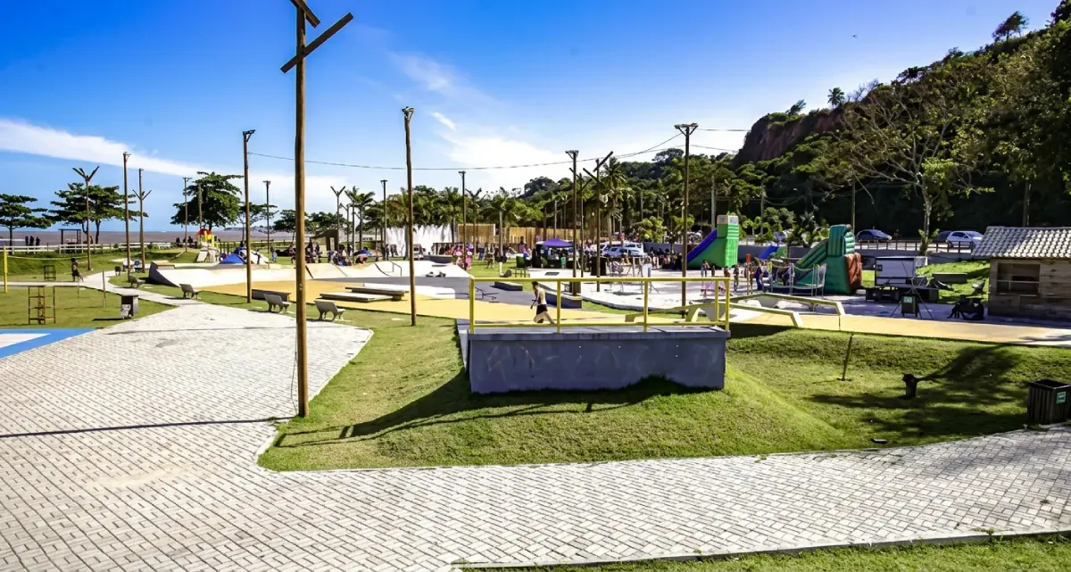 Praça e parque de lazer em Porto Seguro com pavimentação de paralelepípedos, pista de skate, luminárias de madeira e área verde ao lado da falésia e do mar.